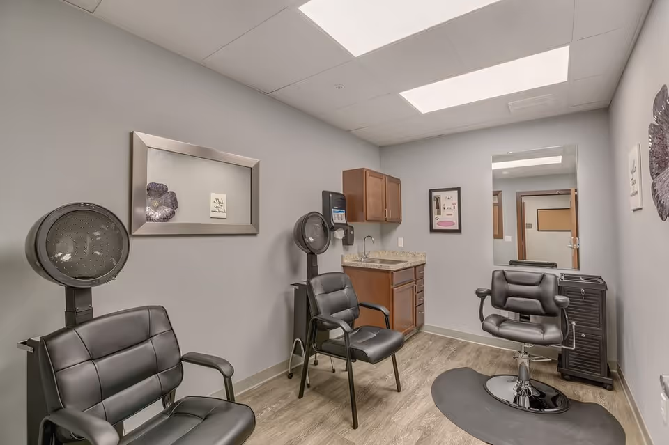 A small salon room with two black salon chairs, one with a hair dryer hood attached, a third black swivel salon chair on a mat, a wooden cabinet with a sink, a wall-mounted soap dispenser, a large mirror, and framed artwork on the light gray walls.