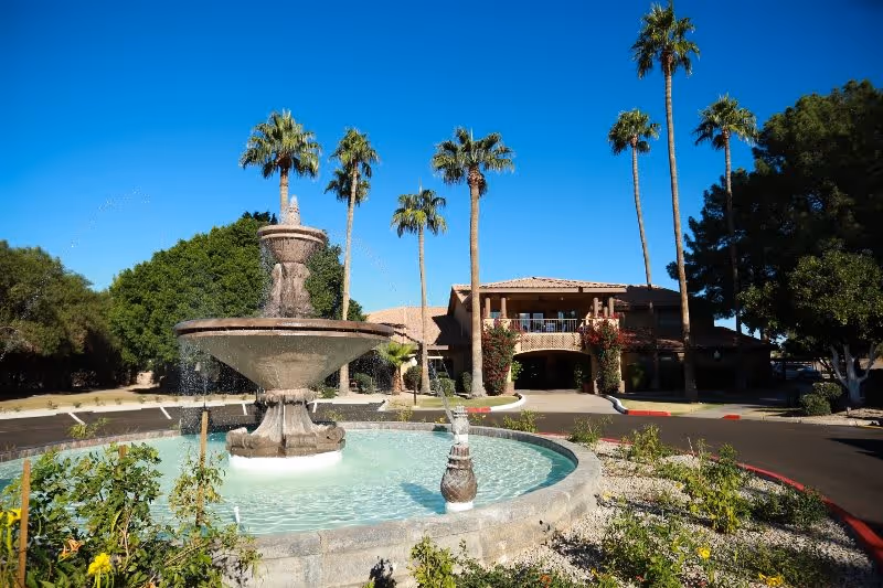 Outdoor view of The Fountains at Lake Pleasant facility featuring a large stone water fountain in the foreground surrounded by plants and a circular driveway. Tall palm trees and other greenery are visible around the building under a clear blue sky.