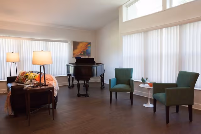 A bright living room with large windows covered by vertical blinds. The room features a black grand piano, a dark sofa with colorful cushions and a throw blanket, two green armchairs, a small round white table with decorative items, and two table lamps on a wooden side table.