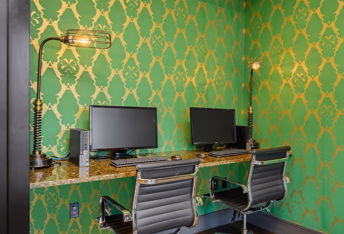 A small computer workstation area with two desktop computers, two black office chairs, and two vintage-style desk lamps. The wall behind the workstations is decorated with green and gold patterned wallpaper.