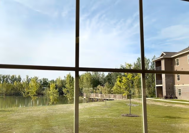 View through a window of a grassy lawn with a small dock on a pond, trees, and a multi-story building to the right.