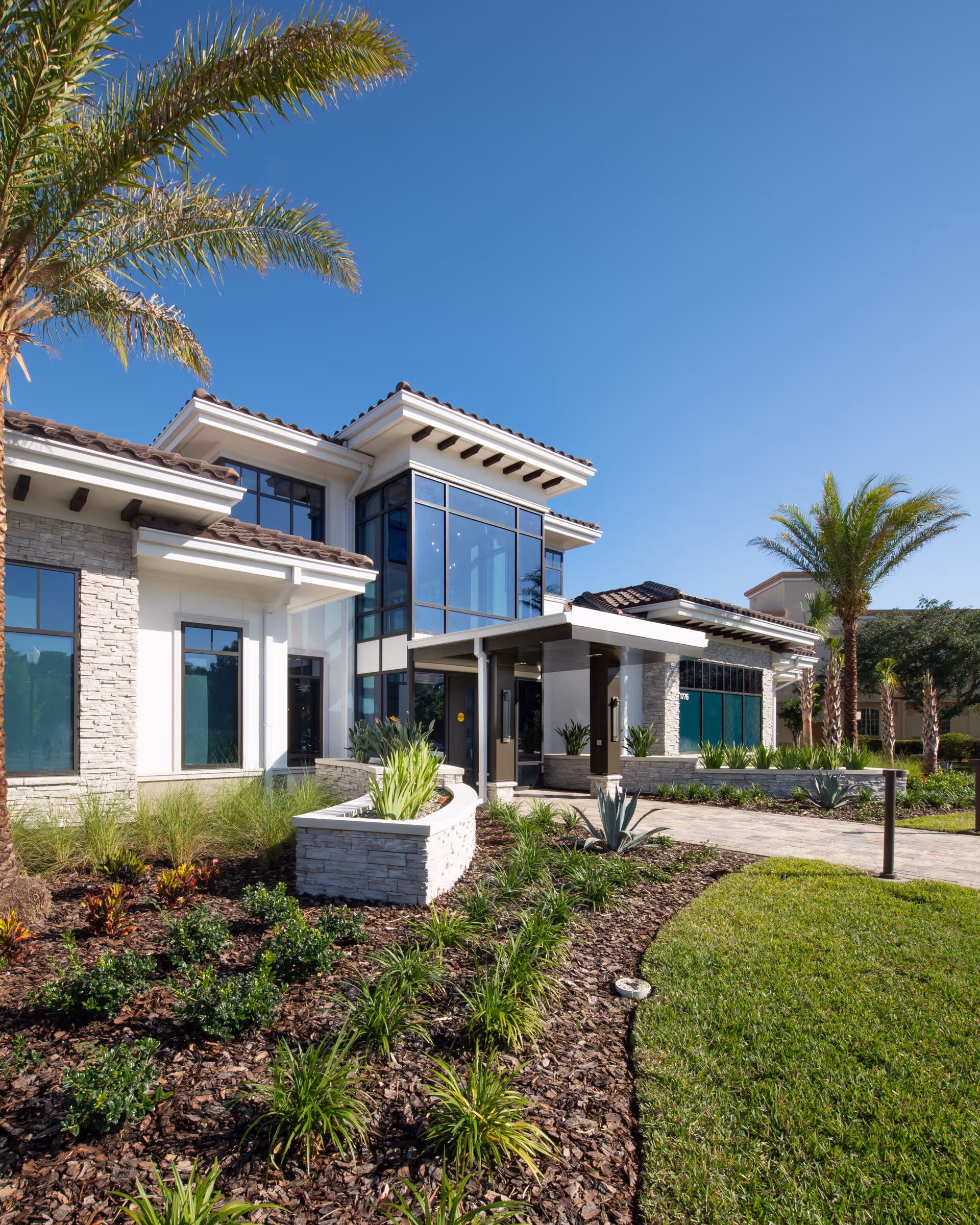 Exterior view of The Mayflower at Winter Park building with modern architecture, large windows, stone and stucco facade, surrounded by landscaped garden beds and palm trees under a clear blue sky.