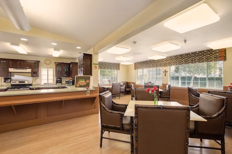 A bright dining room area in a senior living facility with multiple tables and cushioned chairs. The room has large windows with patterned valances allowing natural light to fill the space. Adjacent to the dining area is a kitchen with wooden cabinetry, a stove, and a countertop with a small vase of flowers and a framed picture.
