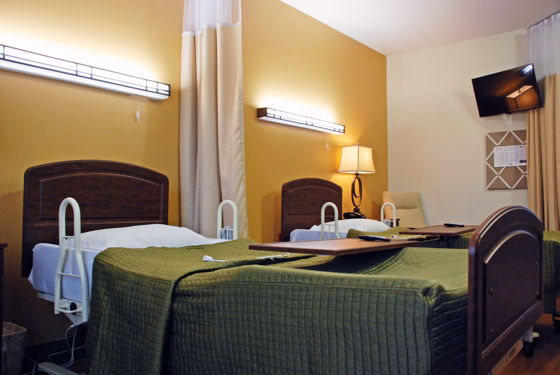 A room with two hospital-style beds with green bedspreads and wooden headboards. Each bed has a white safety rail and an overbed table. The walls are painted yellow and white, with two horizontal wall lights above the beds. A lamp and a chair are visible in the corner, along with a wall-mounted TV and a bulletin board.