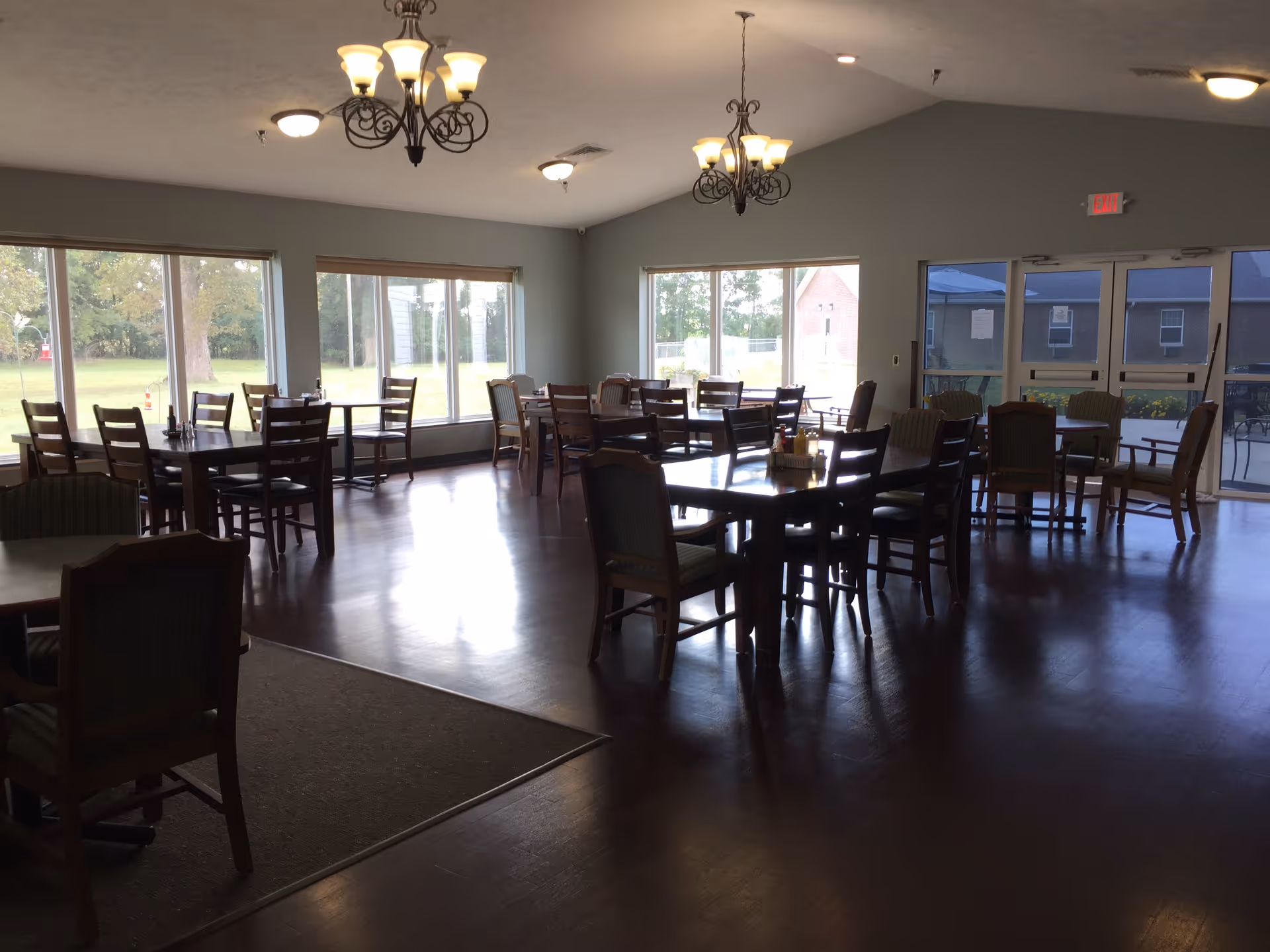A spacious dining room with multiple wooden tables and chairs arranged neatly. Large windows allow natural light to fill the room, offering a view of green outdoor space. Two chandeliers hang from the ceiling, and double glass doors lead outside.