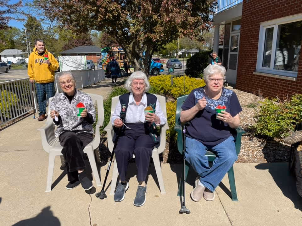 Three elderly women sitting outdoors on plastic chairs enjoying colorful shaved ice treats on a sunny day. Behind them, a man in a yellow Iowa hoodie walks along a sidewalk near a railing, with trees, bushes, and a brick building visible in the background.