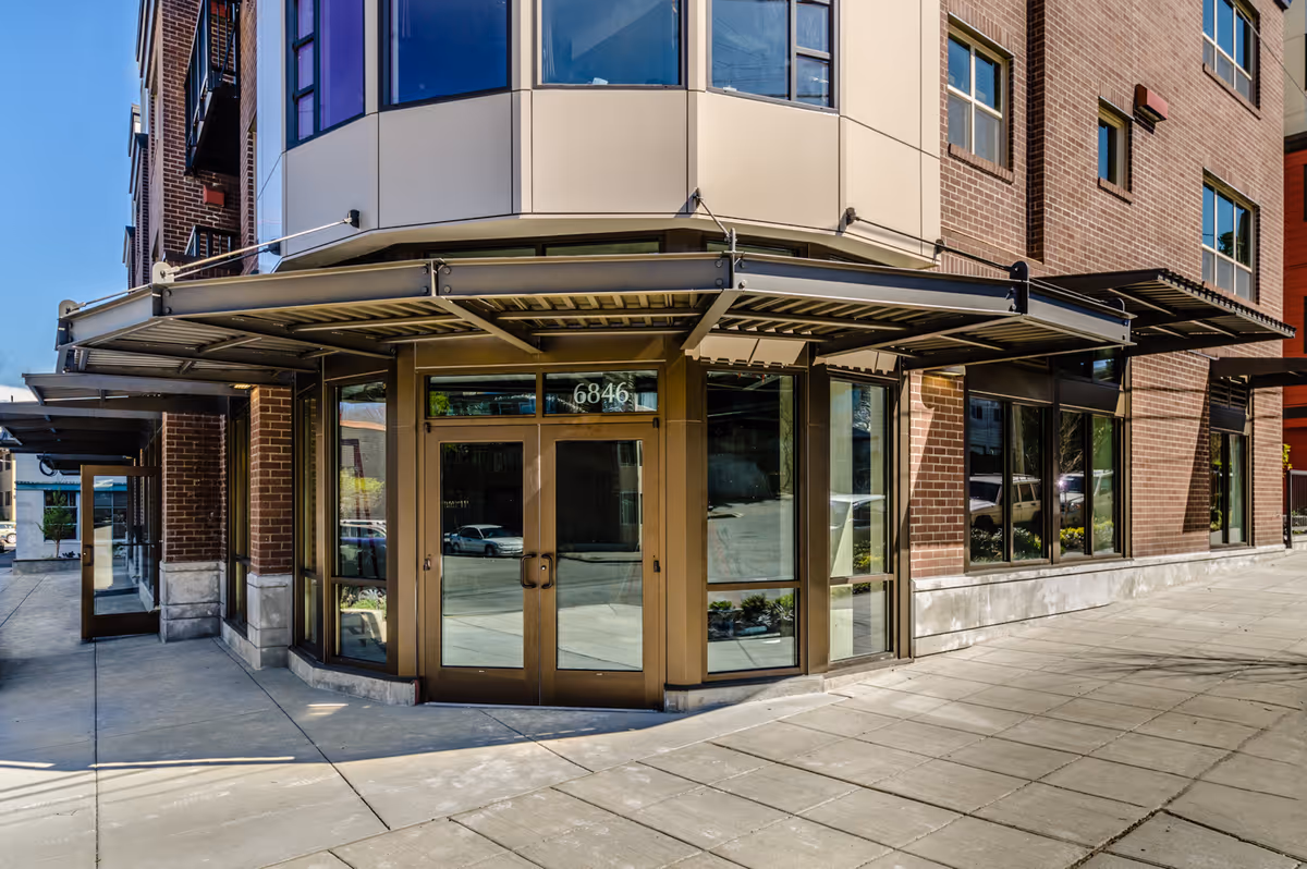 Street-level view of a brick building entrance with glass double doors numbered 6846 and metal awnings.