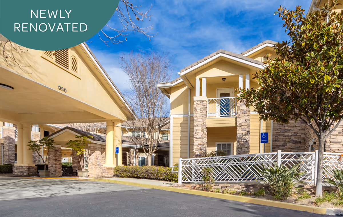 Exterior front entrance of a yellow senior living building with a covered driveway, stone columns, landscaping, and a 'NEWLY RENOVATED' banner.