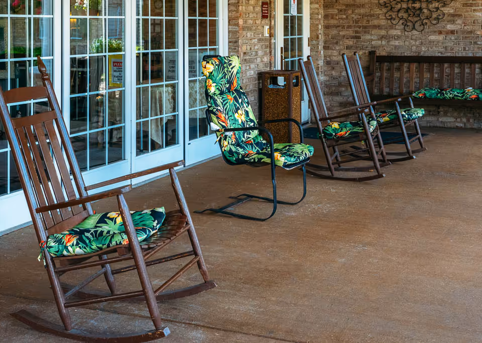 A covered outdoor patio area with several wooden rocking chairs and one metal chair, all with green floral cushions. The patio has a brown floor and brick walls, with large glass doors in the background.