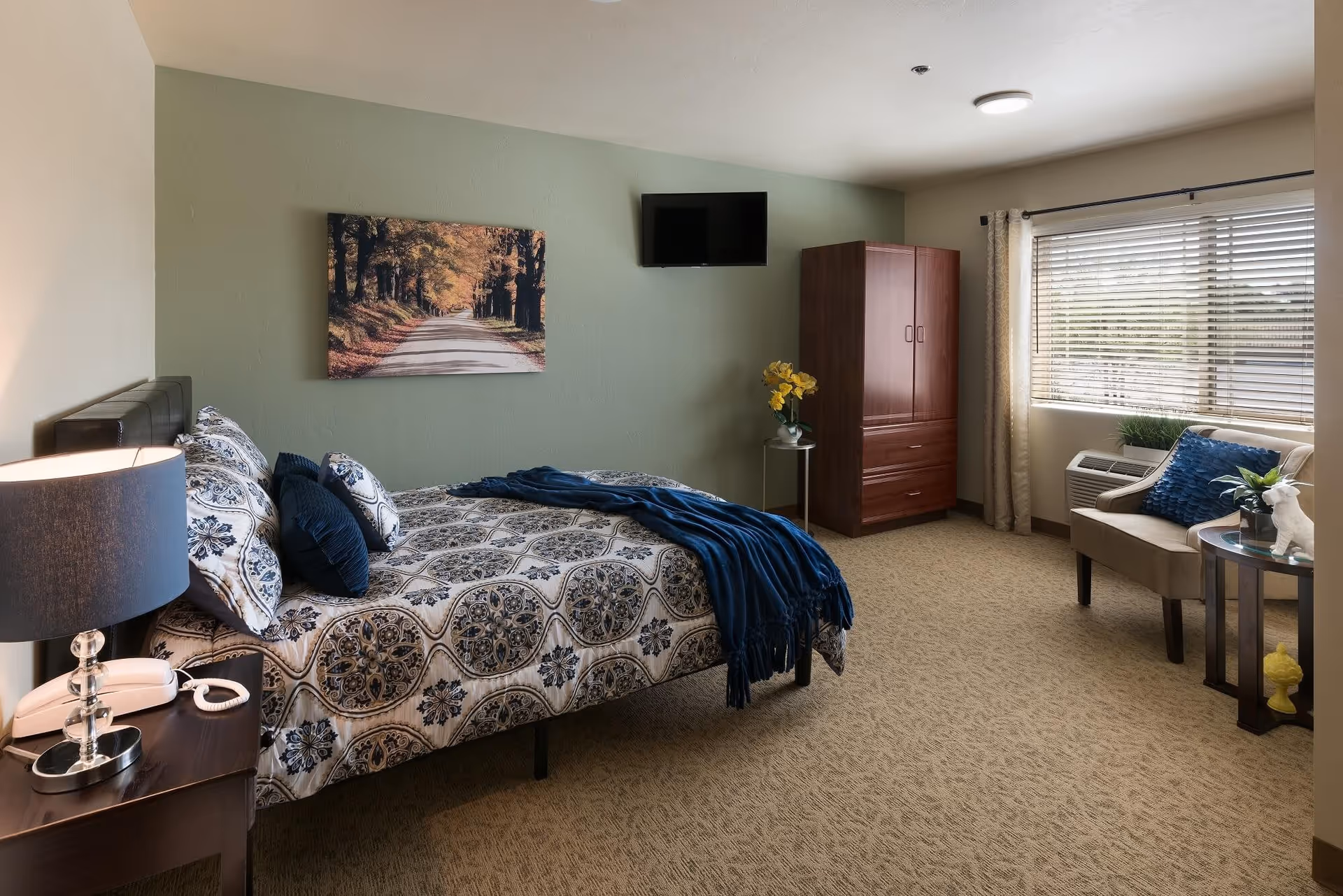 Sunlit furnished bedroom with a patterned bed, armchair and side tables, a wardrobe, wall art, and a window with blinds.