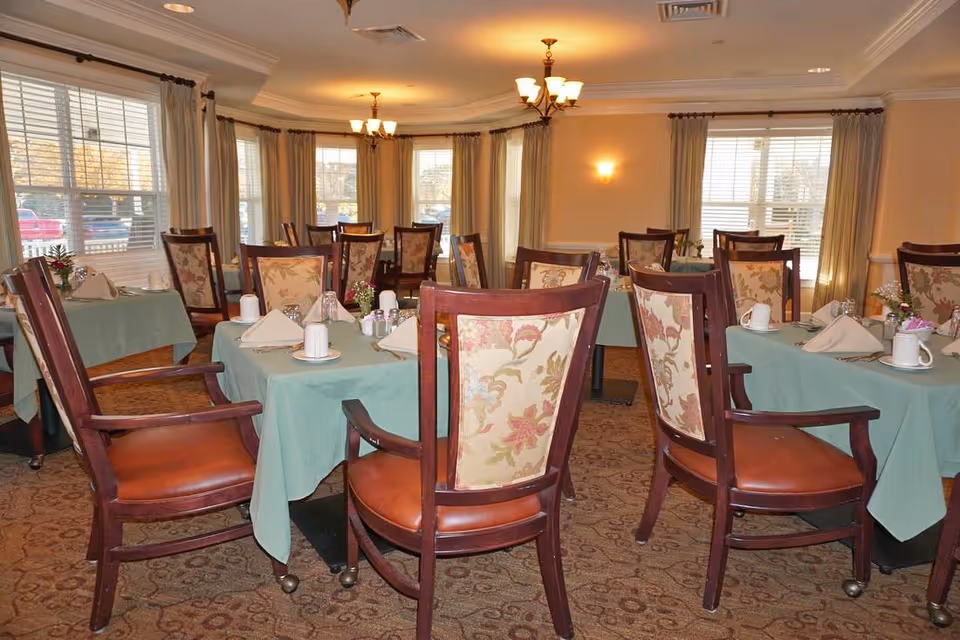 A dining room with multiple tables covered in green tablecloths, each set with white napkins, cups, and small flower arrangements. The room has large windows with beige curtains, patterned carpet, and wooden chairs with floral upholstery on the backrests.