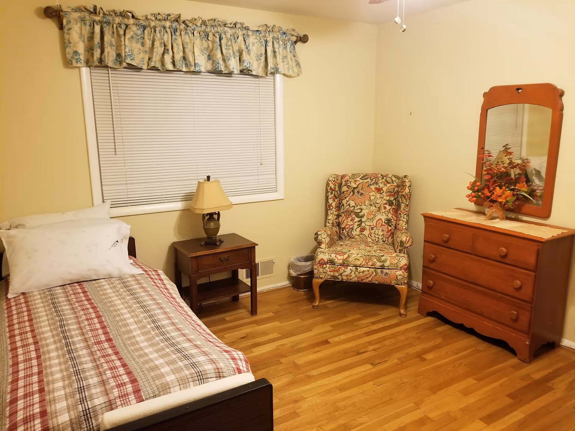 A small bedroom with a single bed covered in a plaid bedspread, a wooden nightstand with a lamp, a floral upholstered armchair, and a wooden dresser with a mirror and a floral arrangement on top. The room has hardwood floors, beige walls, and a window with closed blinds and a floral valance.