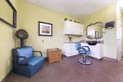 Interior view of a salon area in a nursing home with a blue salon chair in front of a mirror and sink, a blue armchair, a wooden side table, and white cabinets mounted on a light green wall.