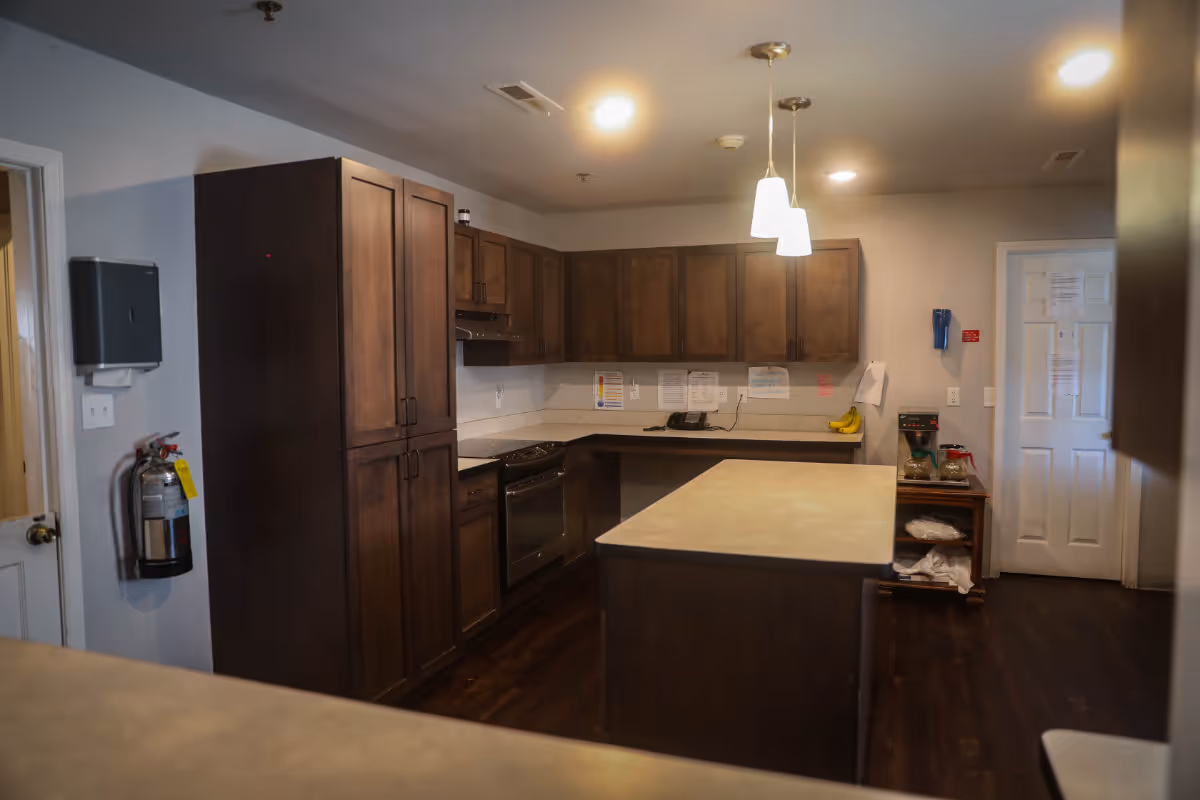 Interior view of a kitchen in Madison Heights Memory Care Community featuring dark wood cabinets, a stove, an island countertop, overhead pendant lights, a fire extinguisher mounted on the wall, and a small table with a coffee maker and bananas.