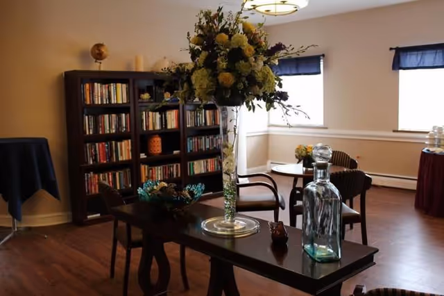 A cozy interior room with wooden flooring featuring a tall vase with a large floral arrangement on a dark wooden table. In the background, there is a bookshelf filled with books, a small round table with chairs, and windows with dark blue curtains letting in natural light.