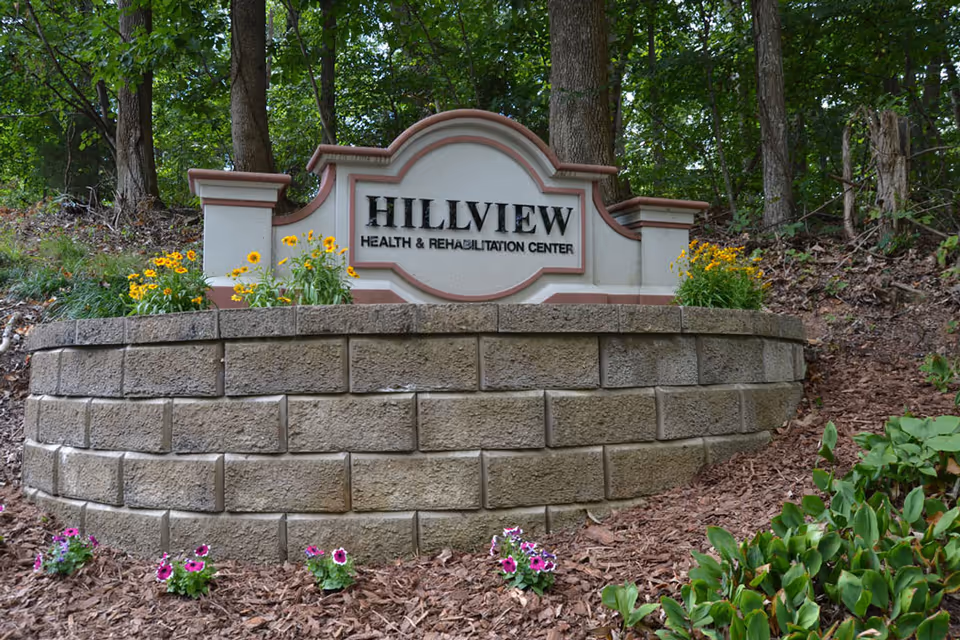 A stone sign for Hillview Health & Rehabilitation Center surrounded by flowers and greenery, with trees in the background.