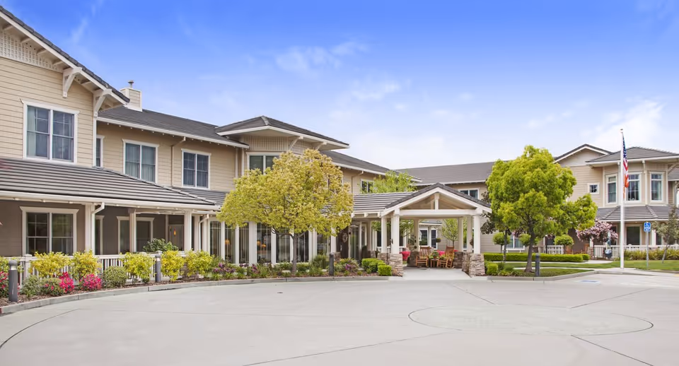 Exterior view of a two-story senior living facility building with beige siding and a covered entrance. There are trees, shrubs, and flowers planted around the building, and an American flag is visible on a flagpole near the parking area.