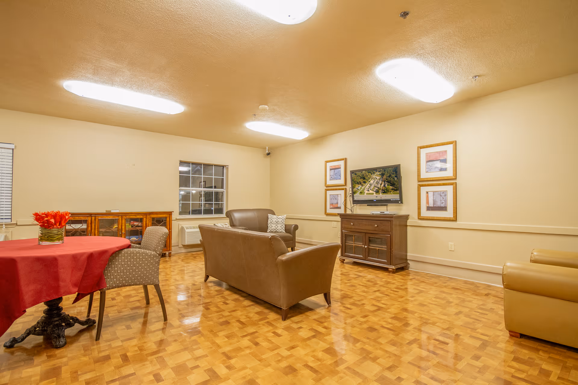 A well-lit living room with wooden parquet flooring, beige walls, and a beige ceiling. The room features a brown leather sofa and armchair facing a flat-screen TV mounted on the wall above a wooden cabinet. There are framed pictures on the wall around the TV. A round table with a red tablecloth and a vase of red flowers is positioned to the left, accompanied by two upholstered chairs. A window with blinds and a wooden sideboard are visible in the background.