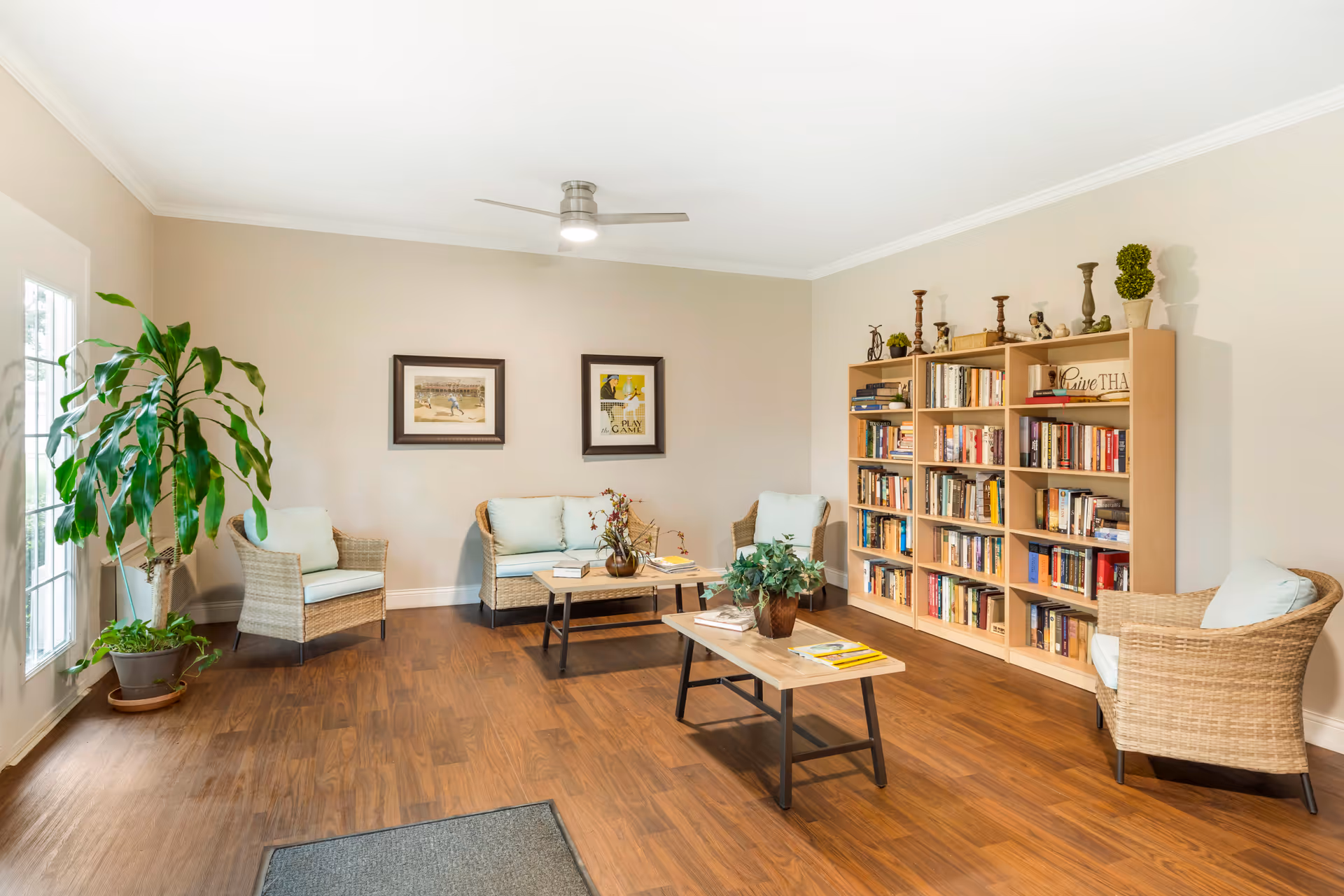 Cozy sunlit common room with wicker seating around coffee tables, a large bookcase, framed art, and potted plants.