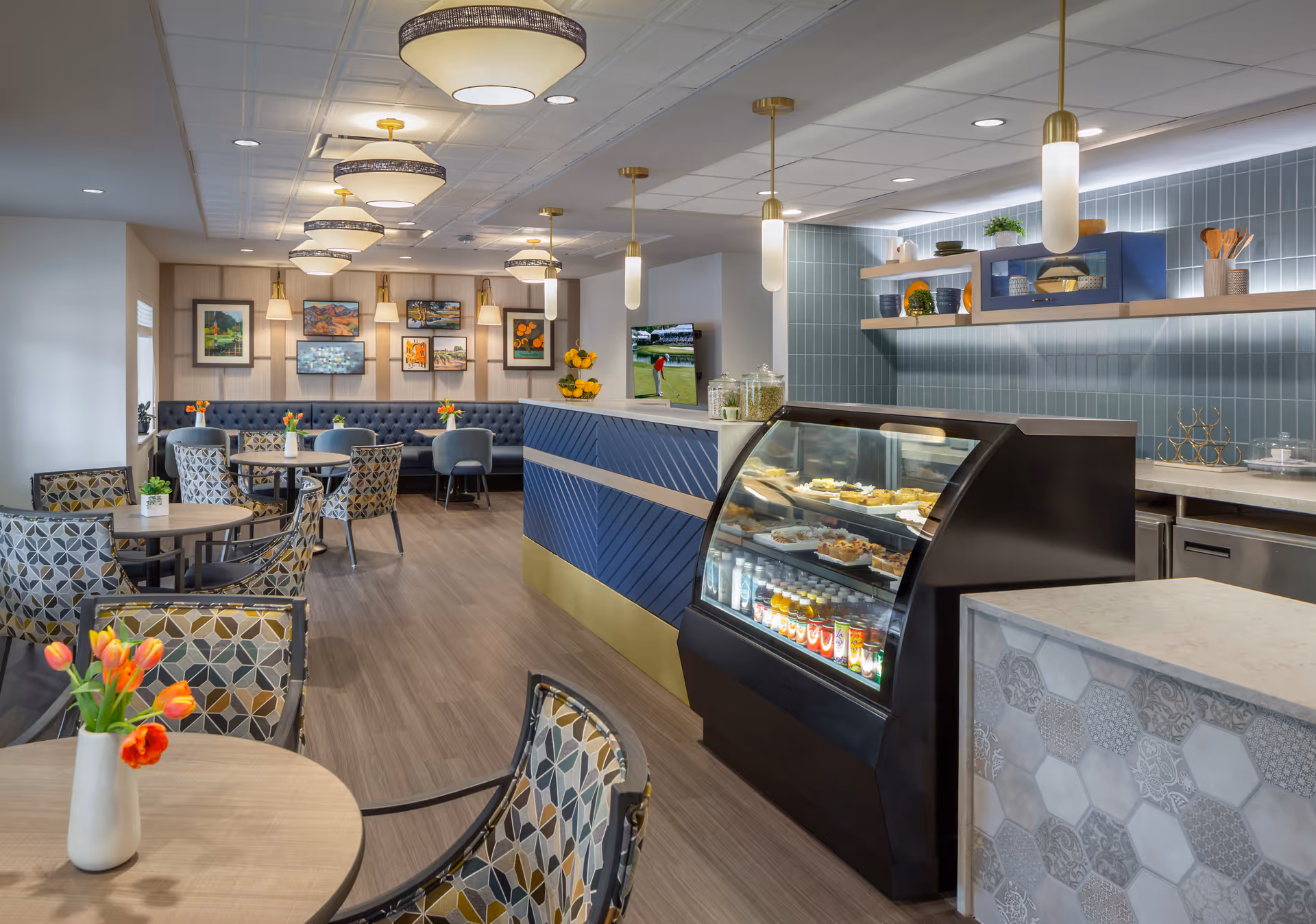 A modern dining area with patterned chairs and round tables adorned with small vases of orange tulips. The room features a blue and gold service counter with a glass display case containing beverages and pastries. The back wall has blue tile and wooden shelves with decorative items, and there is a TV mounted on the wall showing a golf game. The ceiling has multiple pendant lights and the far wall is decorated with framed artwork above a blue cushioned bench.