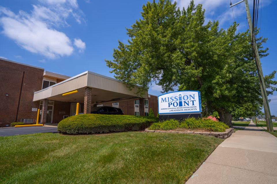 Front entrance of Mission Point Nursing & Physical Rehabilitation Center with a covered drop-off, sign, and landscaped lawn.
