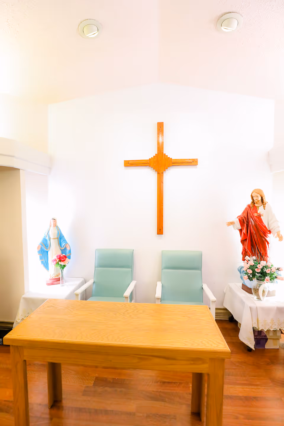 Small chapel-like interior with a wooden cross on the wall, two mint chairs, a wooden table and statues of Mary and Jesus.