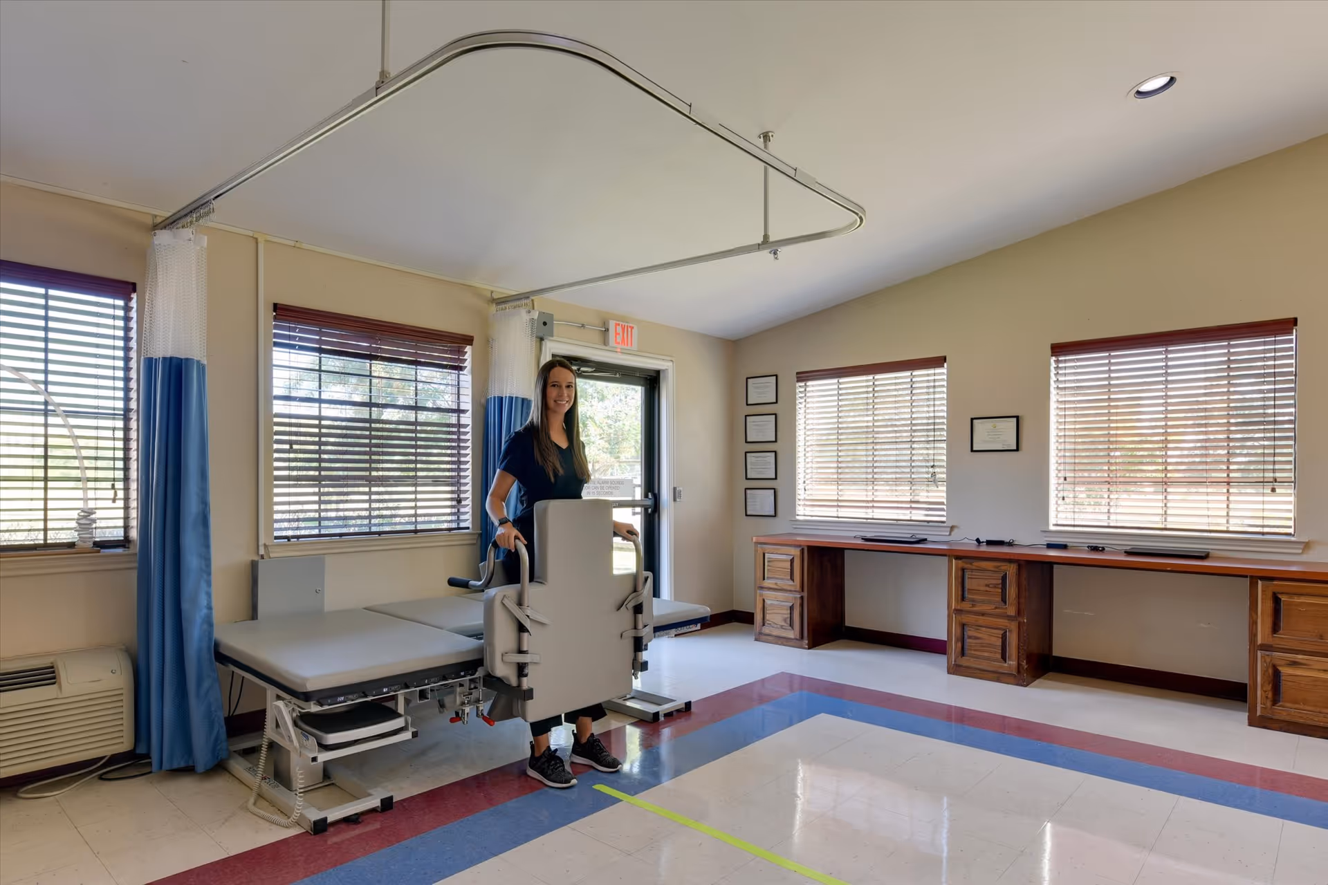 A woman standing next to a medical examination table in a well-lit room with large windows covered by blinds. The room has a tiled floor with red and blue borders, a long wooden desk with multiple drawers along one wall, and blue and white privacy curtains hanging from a ceiling track.