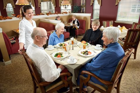 Four elderly people sitting around a dining table in a restaurant-style setting, enjoying a meal with plates of food and drinks. A smiling waitress stands beside the table. In the background, another elderly person is seated at a separate table. The room has large windows with blinds and warm lighting.