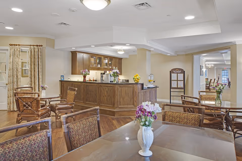 A bright and inviting common area in a senior living facility featuring a wooden reception or serving counter with flowers and a bowl of lemons on top. Surrounding the counter are several tables and chairs with patterned upholstery, each table decorated with a vase of flowers. The room has hardwood flooring, beige walls, and recessed lighting in the ceiling. A signboard with the Sunrise logo is visible near a doorway leading to another room with more seating.