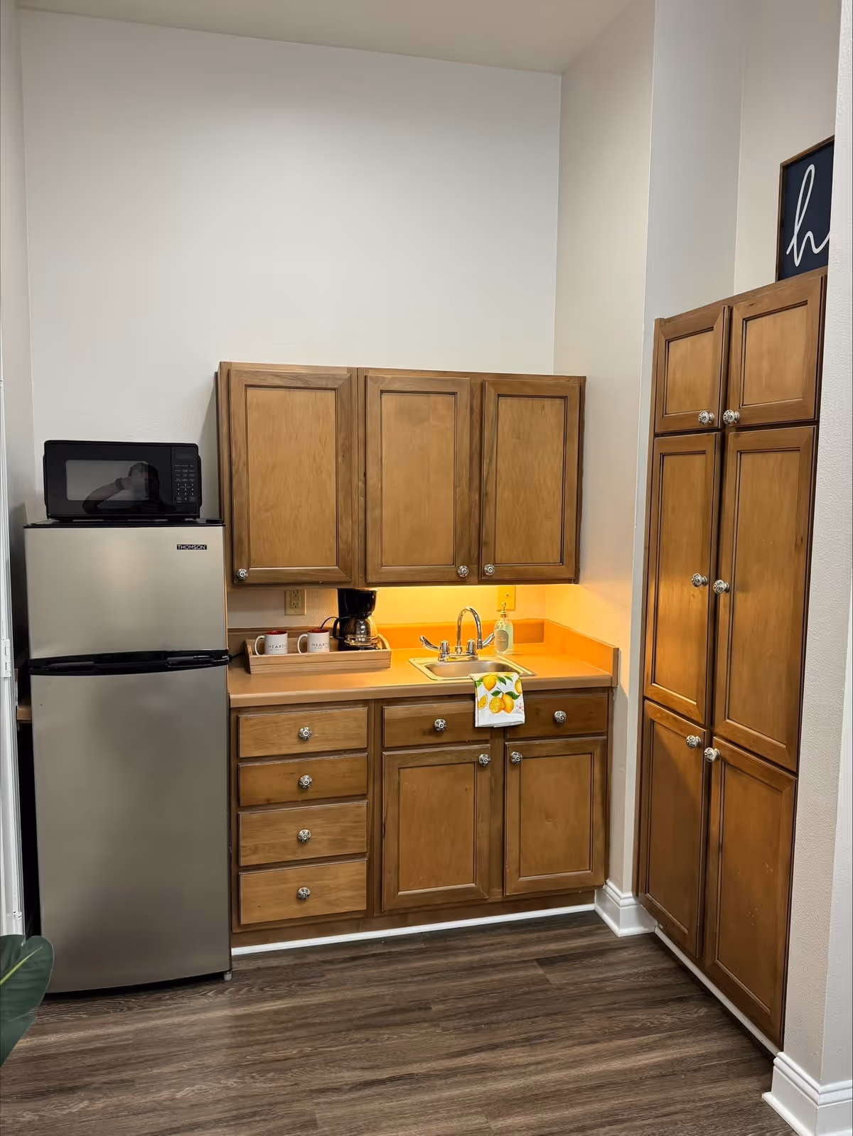 A small kitchen area with wooden cabinets, a stainless steel refrigerator with a microwave on top, a countertop with a coffee maker, two mugs, a sink with a faucet, and a lemon-themed hand towel hanging from the cabinet handle. The floor is wood, and the walls are white.
