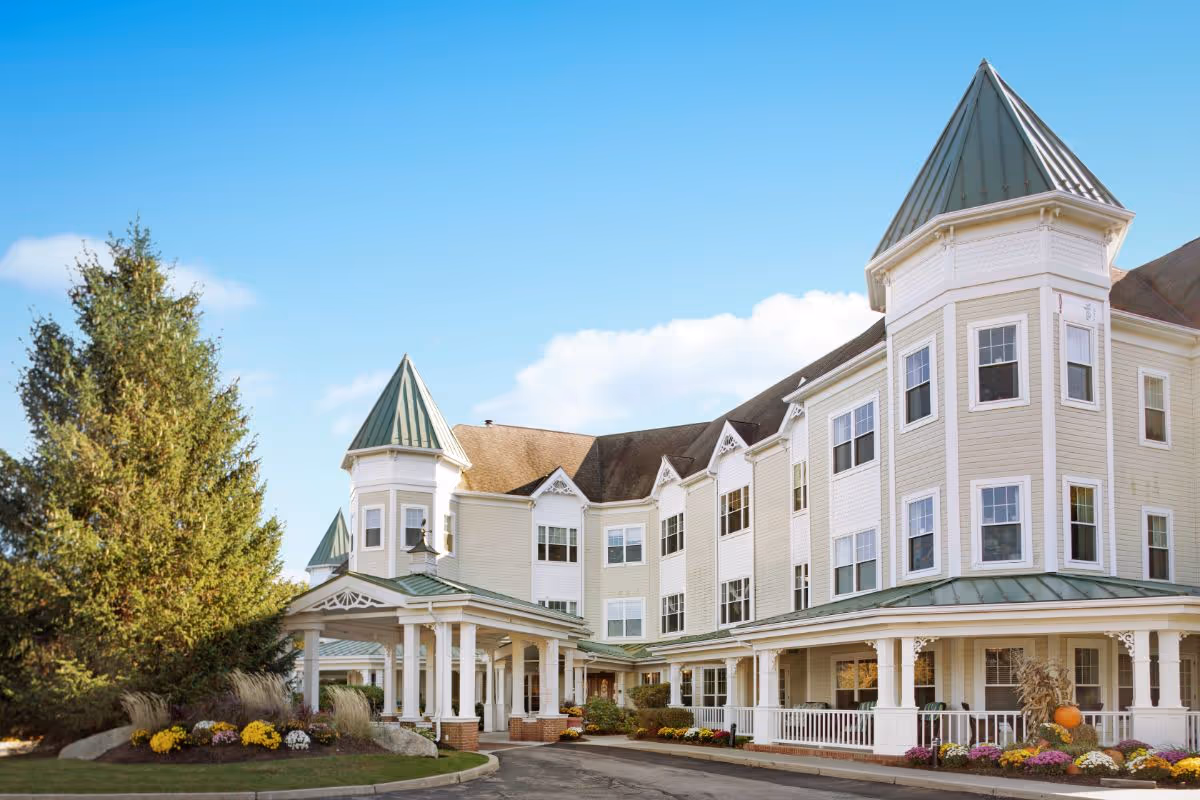 Front exterior of a large multi-story senior living building with turreted rooflines, a covered entrance, and landscaped flower beds under a blue sky.