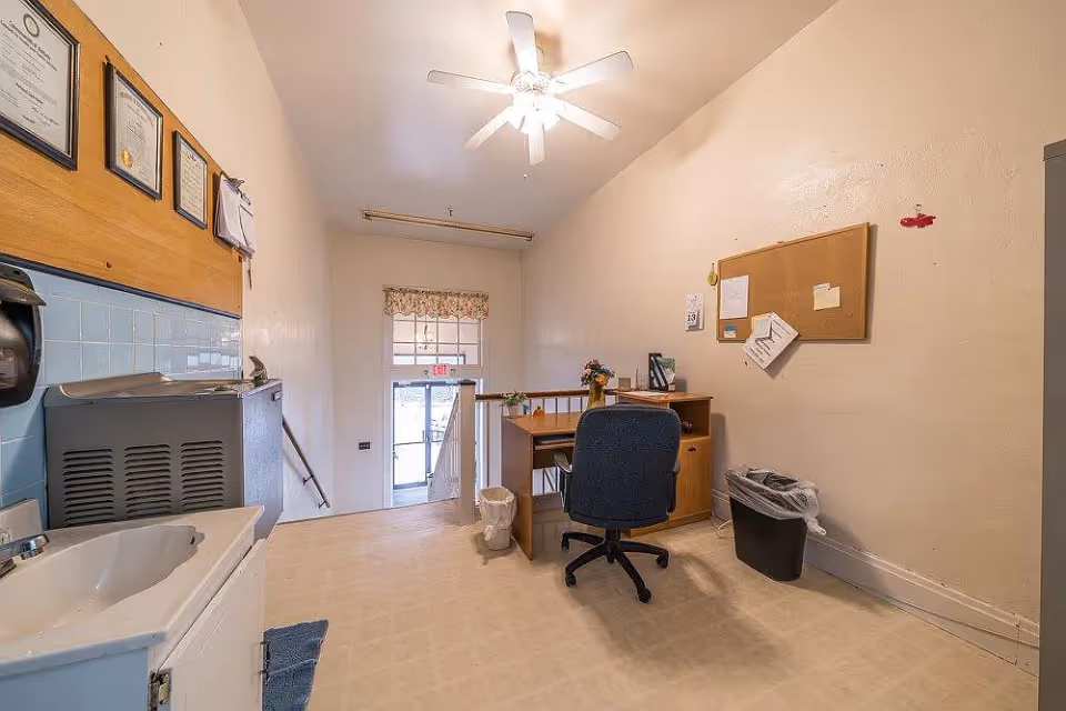 Small office area with a desk, office chair, cork bulletin board with papers pinned, trash can, and a sink with a paper towel dispenser mounted on the wall. Certificates or documents are framed and hung above the sink. A ceiling fan with light is on the ceiling, and a door with a window and an exit sign is visible at the far end.