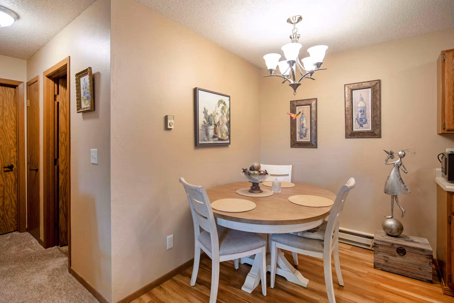 A cozy dining nook with a round wooden table and four chairs under a chandelier, decorated with wall art and a metal figurine.