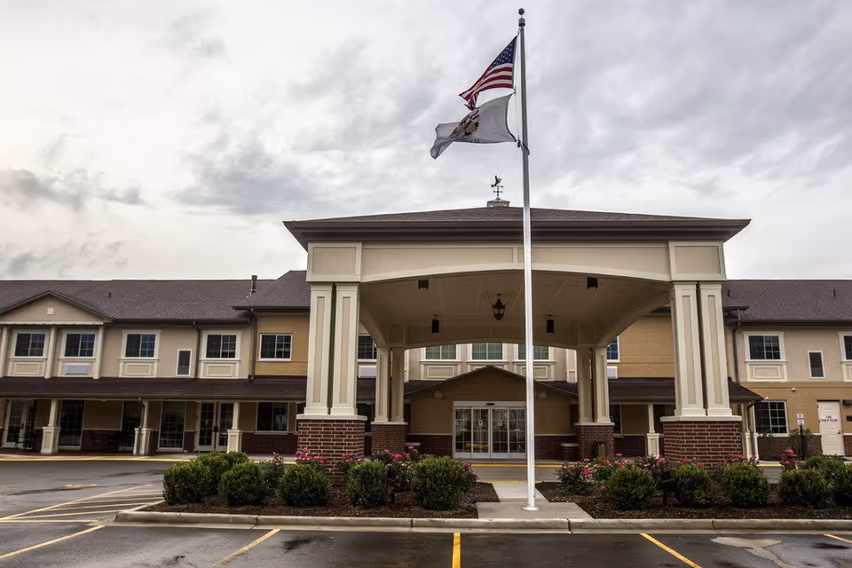 Front exterior view of Cedar Lake Assisted Living and Memory Care building with a covered entrance, American and state flags on a flagpole, and landscaped bushes and flowers in front.