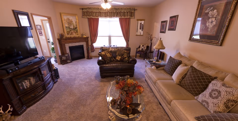 A cozy living room with a beige sofa adorned with patterned pillows, a dark leather armchair, a glass coffee table with a decorative centerpiece, a large flat-screen TV on a wooden stand, a fireplace with a framed painting above it, and a window with red curtains letting in natural light.