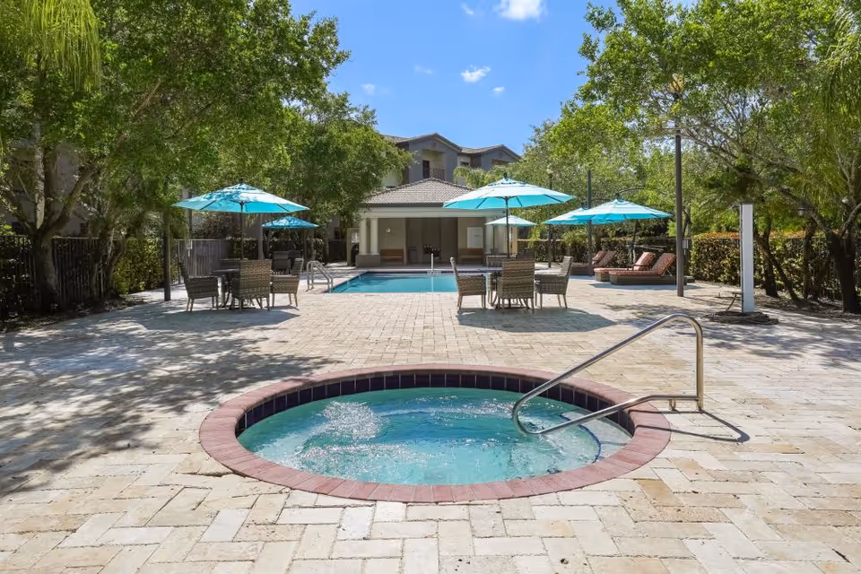 Outdoor pool area at Verena at Delray featuring a circular hot tub in the foreground with bubbling water, a rectangular swimming pool in the background, several tables with turquoise umbrellas, lounge chairs, and surrounded by trees under a clear blue sky.