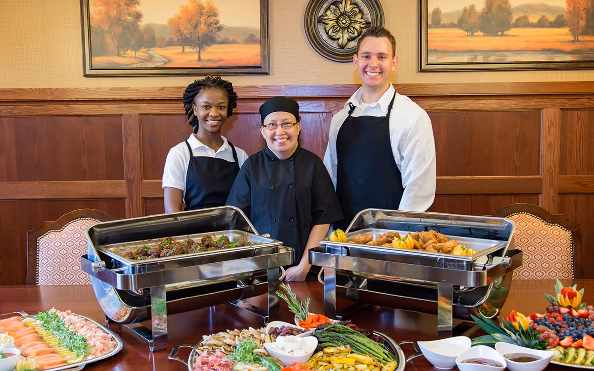 Three catering staff members standing behind a buffet table with trays of food including meat dishes, vegetables, and fruit platters in a dining room with wooden paneling and framed landscape paintings on the wall.
