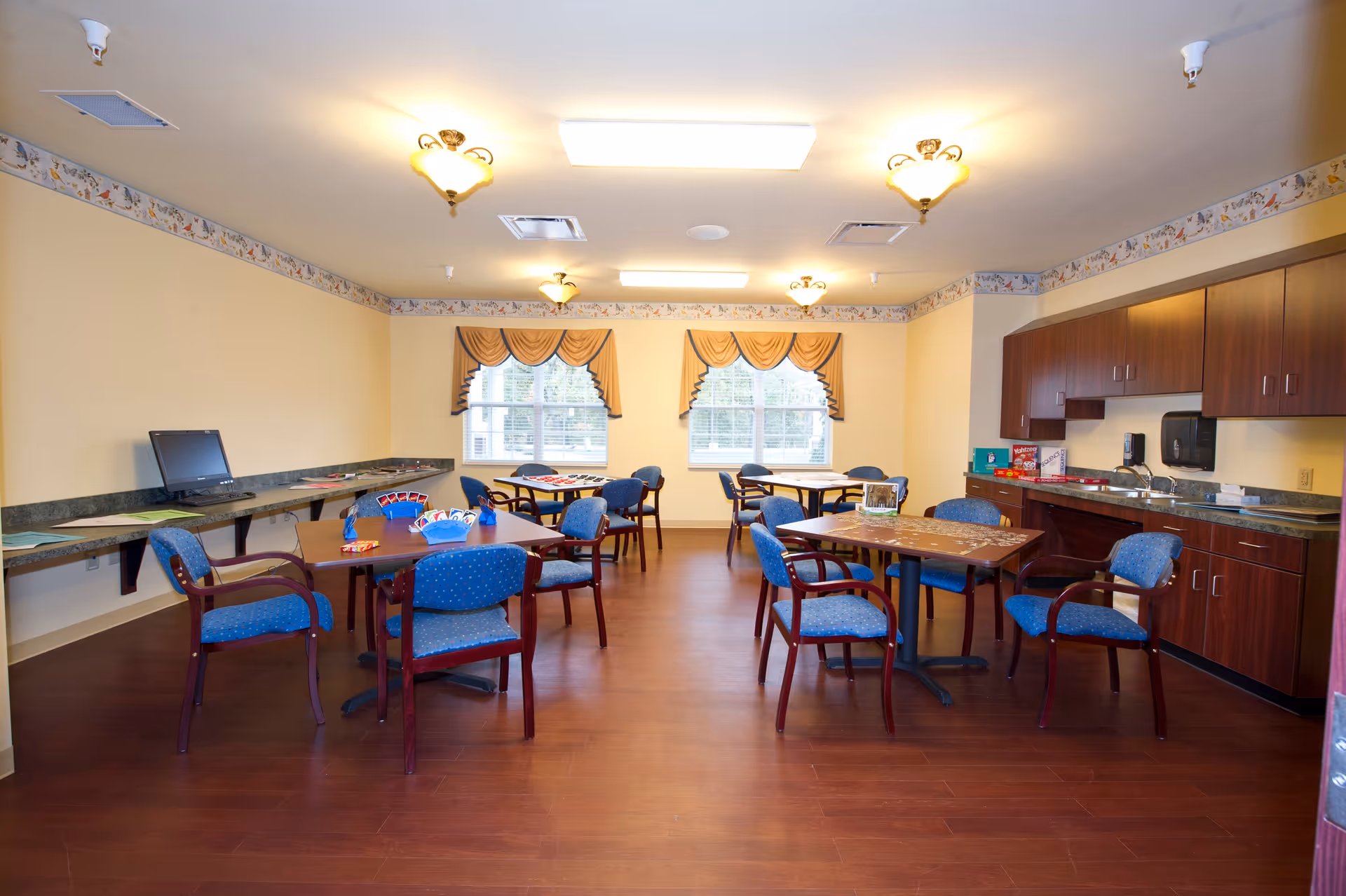 Bright communal activity/dining room with several tables and blue chairs, a kitchenette and a computer station.