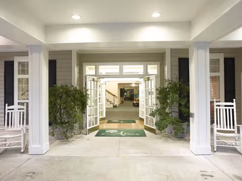 Front entrance of Forest Lake Manor with double doors open, potted plants and rocking chairs.