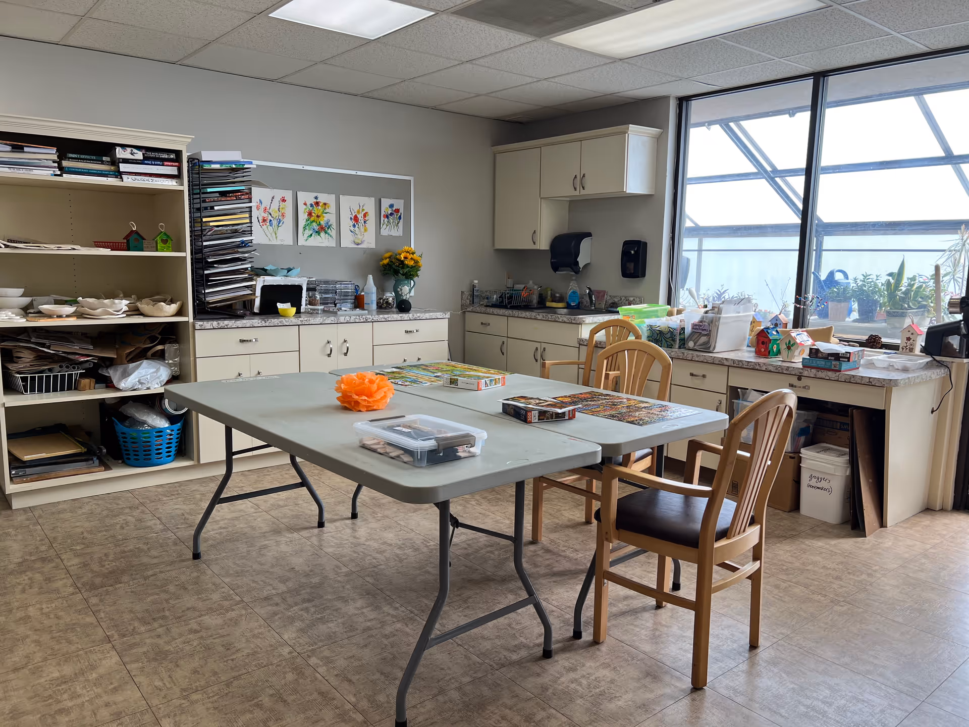A well-lit activity room with a large window showing plants outside. The room contains a folding table with puzzles and craft supplies on it, surrounded by wooden chairs. Cabinets and shelves along the walls hold various art materials, books, and decorations. Colorful flower paintings are displayed on a bulletin board above the counter.