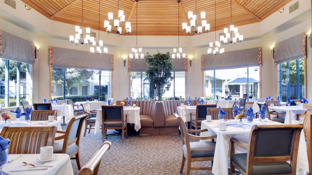 Bright, elegant dining room with a wooden domed ceiling and chandeliers, tables set with white linens and blue napkins, and large windows looking out to greenery.