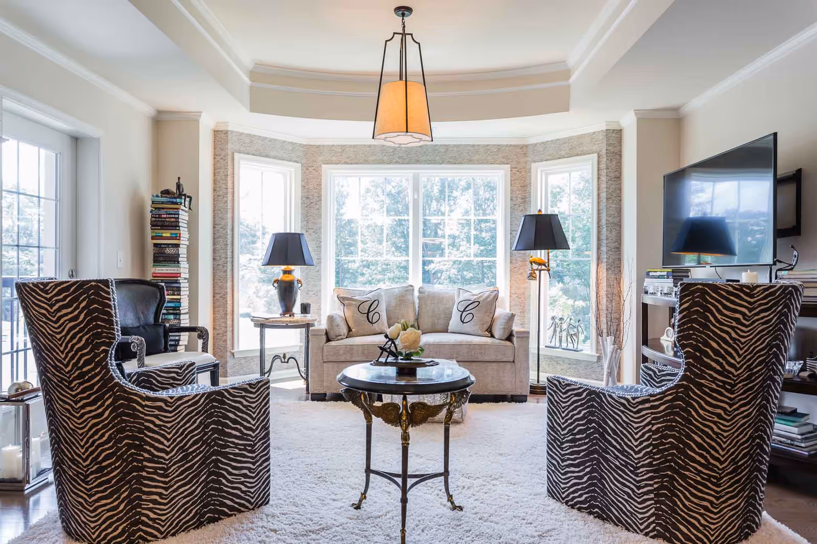 Bright and elegant living room with large windows letting in natural light, featuring a beige sofa with two decorative pillows, two zebra-patterned armchairs, a round coffee table with a flower arrangement, two black lamps on side tables, a flat-screen TV mounted on the wall, and a tall stack of books in the corner.