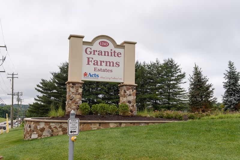 Outdoor view of a large stone and stucco sign for Granite Farms Estates with trees and grass surrounding it under a cloudy sky.