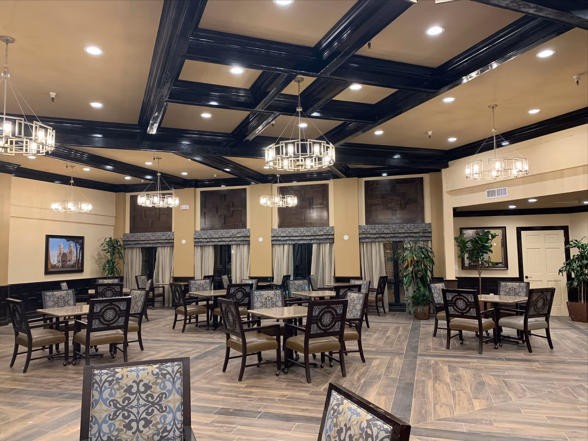 A spacious dining room with multiple tables and chairs arranged neatly. The room features a decorative coffered ceiling with dark wood beams and modern chandeliers. Large windows with patterned valances and curtains line the walls, and there are several potted plants placed around the room. The floor has a wood-like tile design, and the walls are painted in a warm beige tone with dark wood paneling along the lower half.