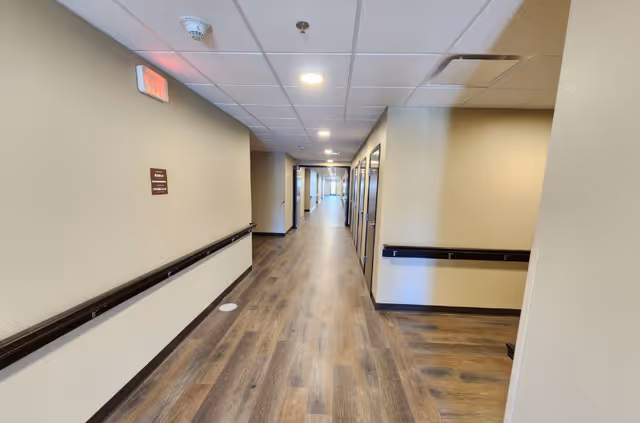 Long, well-lit interior corridor with handrails, wood-look flooring, and multiple doorways in a senior living facility.