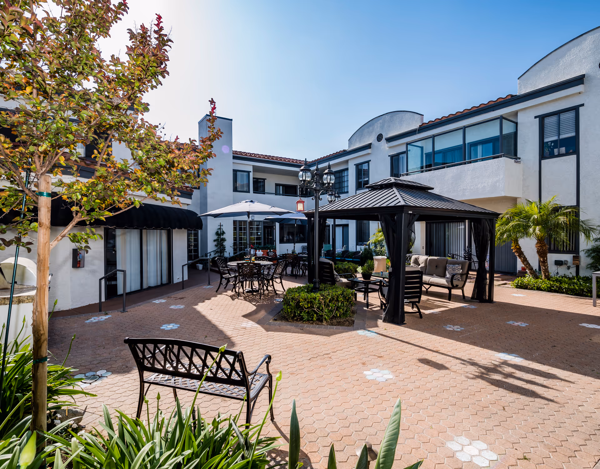 Sunlit courtyard with a gazebo, outdoor seating and tables with umbrellas surrounded by a two-story building.