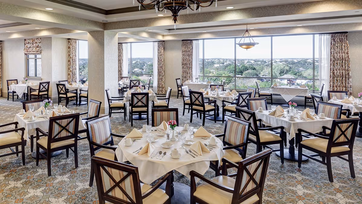 A spacious dining room with multiple tables covered in white tablecloths, each set with beige napkins, cups, glasses, and silverware. The room features large windows with floral curtains, allowing natural light and a view of trees and distant buildings. The chairs have wooden frames with striped cushions, and the floor is covered with a patterned carpet.