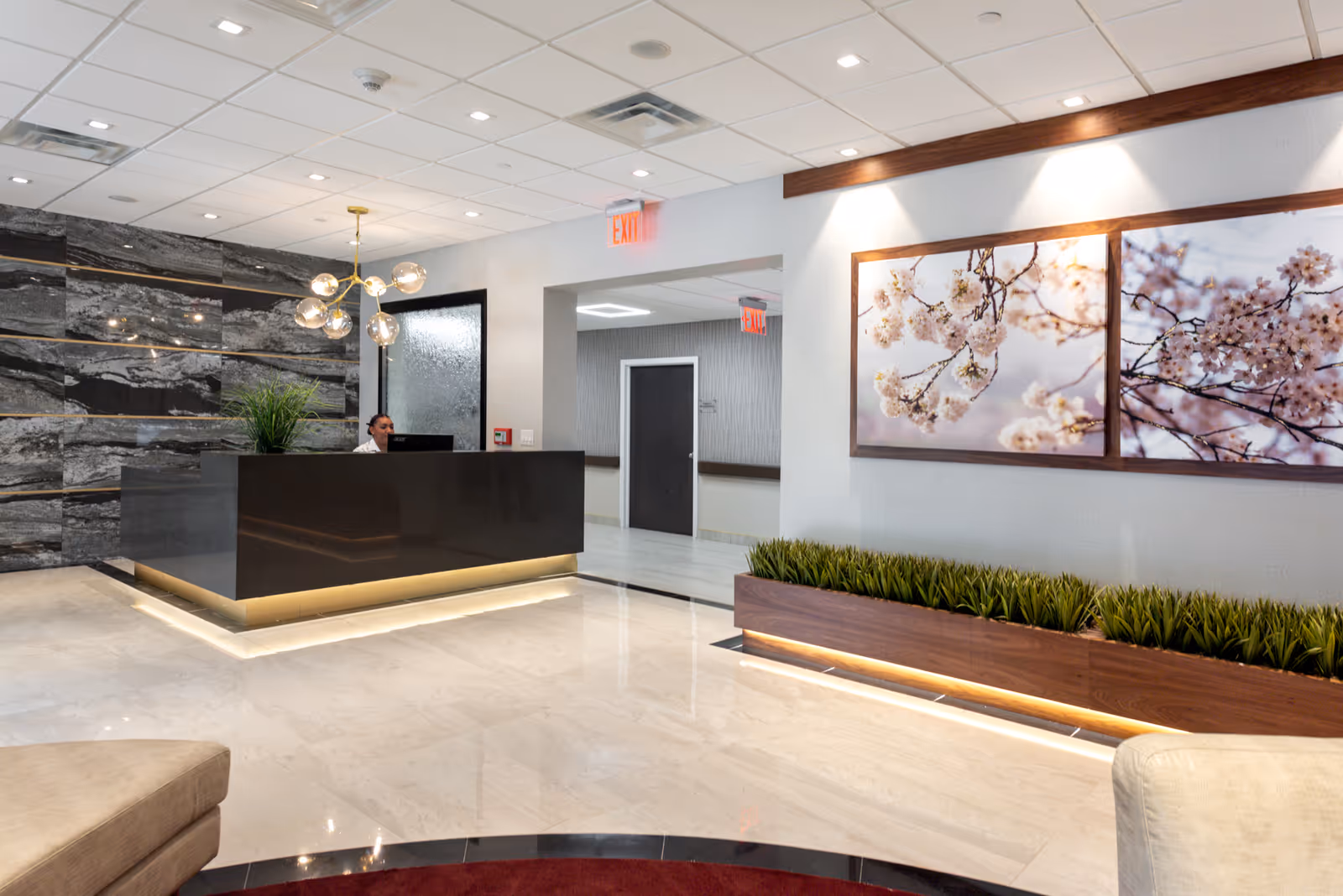 Modern reception area with a dark sleek front desk, a receptionist seated behind it, a decorative wall with black and gray stone tiles, and a contemporary chandelier. The floor is shiny and light-colored, with a planter box containing green plants along the right wall. Two framed pictures of cherry blossoms hang above the planter, and there are exit signs visible in the hallway beyond the reception desk.