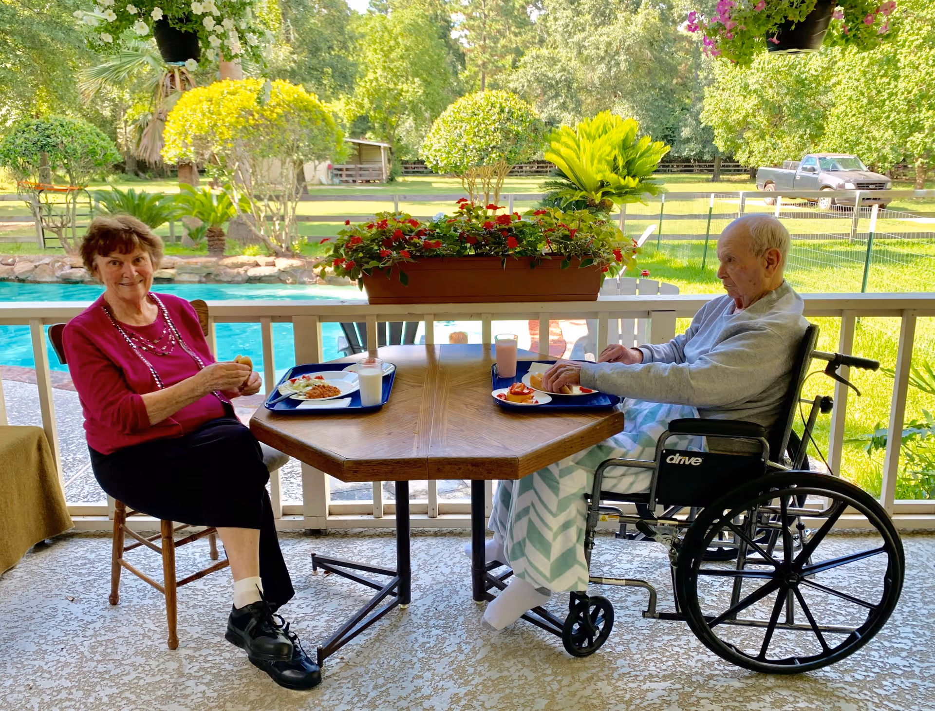Two elderly individuals sitting at a wooden table on a covered patio. One woman is seated on a wooden chair wearing a red top and black pants, smiling at the camera. The other person, a man, is seated in a wheelchair with a blanket over his legs. Both have trays with food and drinks in front of them. Behind them is a swimming pool, green lawn, trees, and a parked pickup truck in the background.