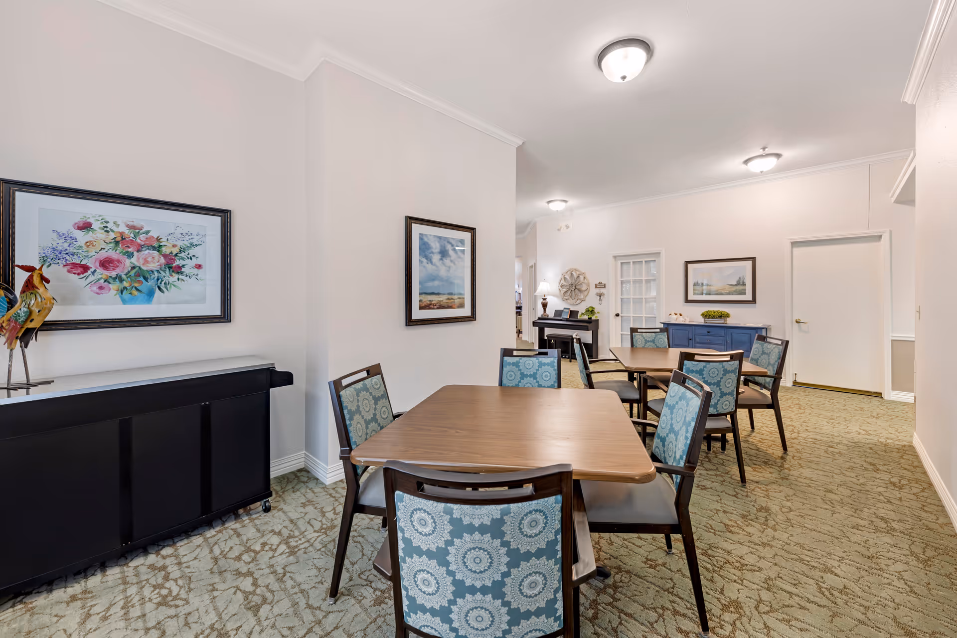 A dining area with two wooden tables surrounded by chairs with blue patterned upholstery. The room has light-colored walls adorned with framed artwork, a carpeted floor, and ceiling lights. There is a black piano against one wall and a blue sideboard with decorative items on the far wall.
